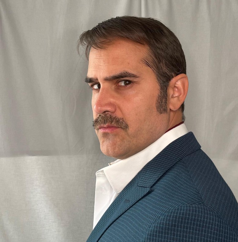 Andreas Barås i blå kavaj och mustasch, karaktärsporträtt.